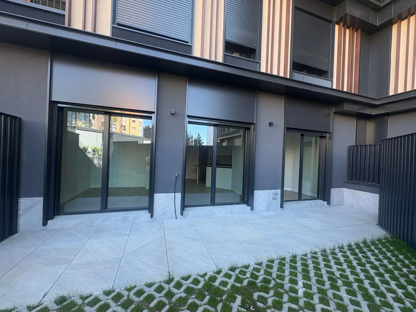 Exterior view of a modern gray building with three sliding glass doors opening onto a tiled patio and grass pavers.
