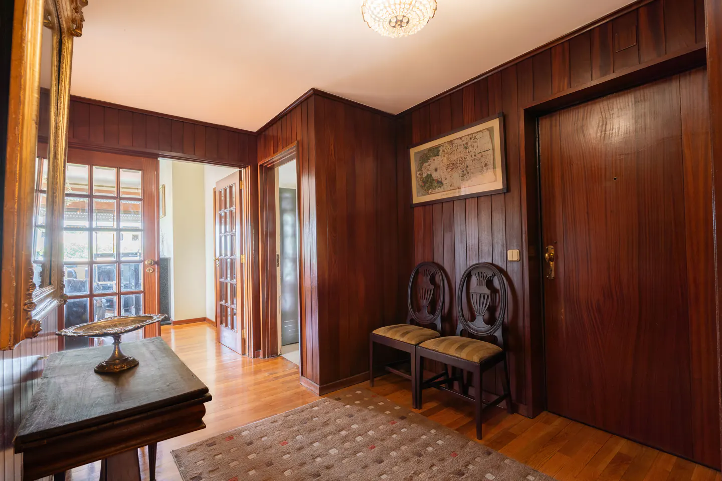 A wood-paneled hallway with a table, chairs, and doors. A rug is on the floor.