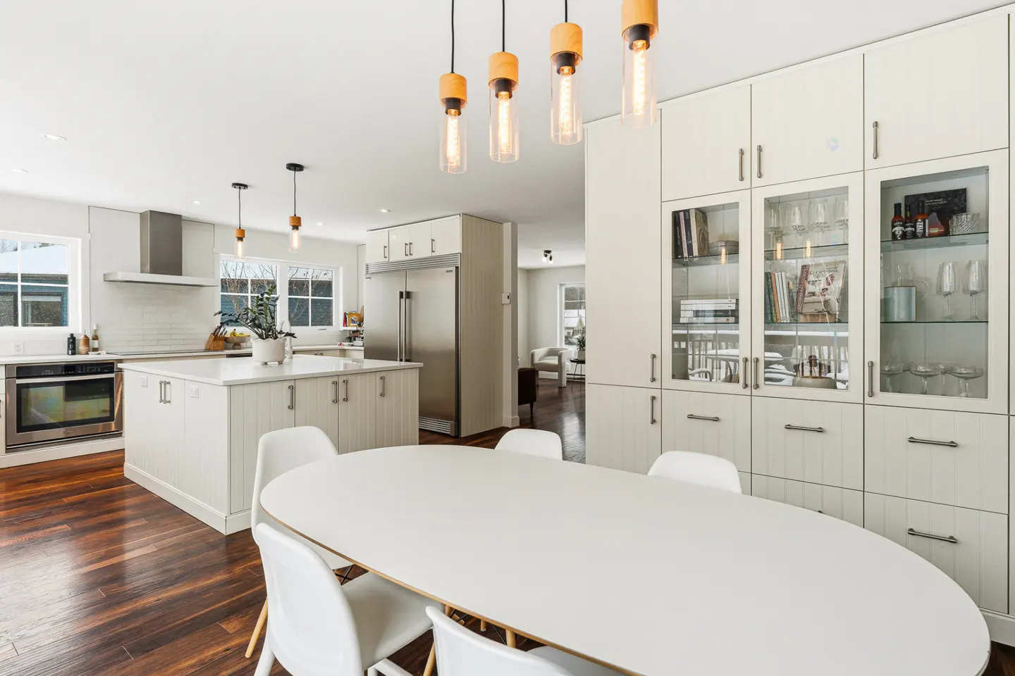 Bright, modern kitchen with white cabinets, stainless steel appliances, and a dark wood floor. A white oval table with chairs sits in the foreground.