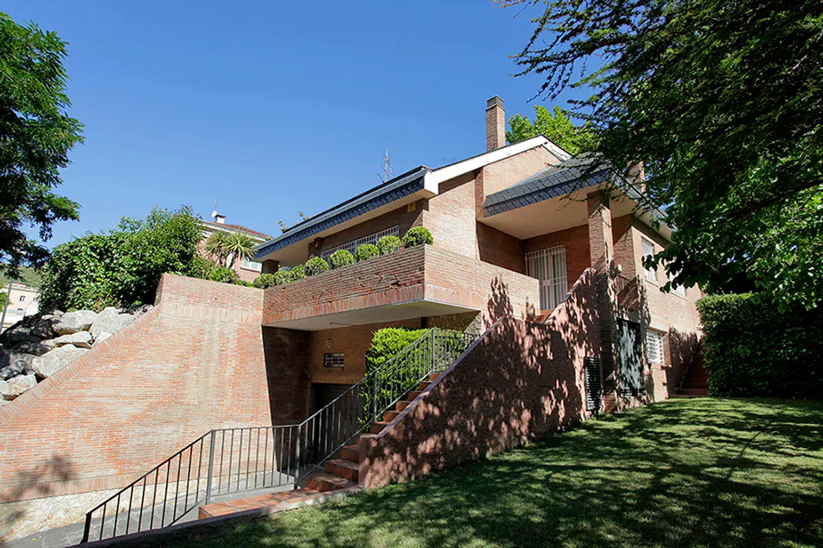 Brick house with a dark roof, a balcony with plants, and a staircase leading to the entrance, surrounded by green trees and grass.