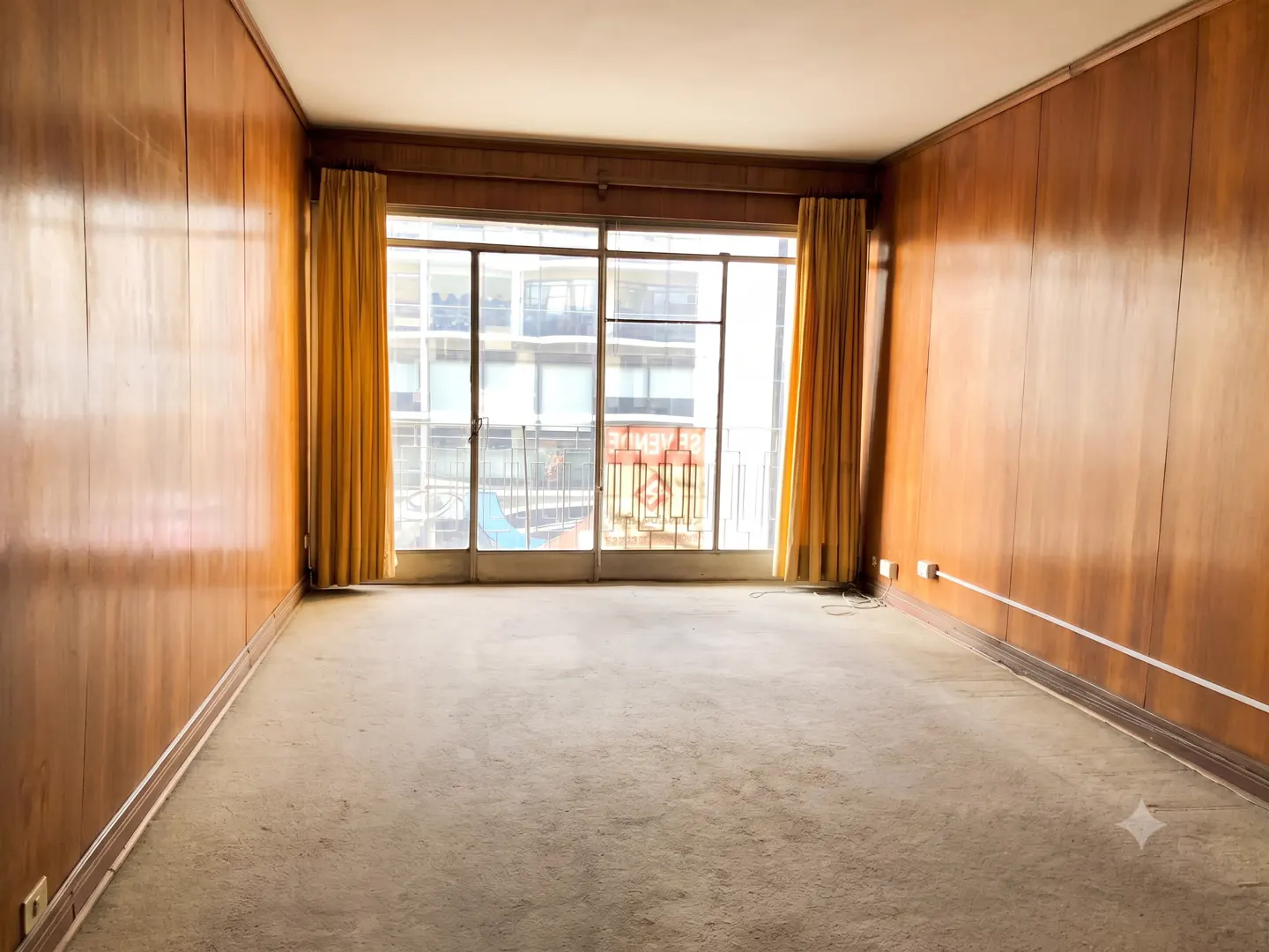 Empty room with wood-paneled walls, beige carpet, and large glass doors with yellow curtains leading to a balcony.