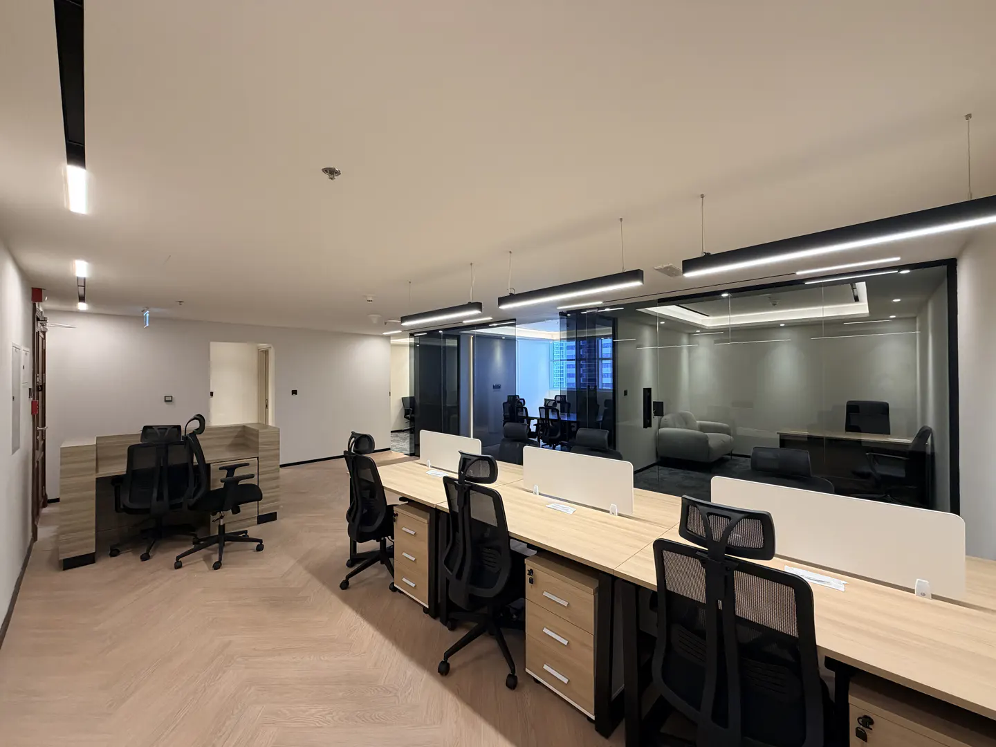 A modern office space with long desks, black chairs, and glass-walled conference rooms. The floor is light wood.