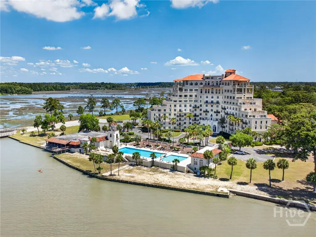 Aerial view of a white, multi-story resort with a red tile roof, a pool, and palm trees, on a riverfront property under a blue sky.
