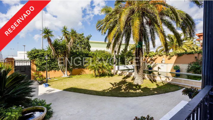 A lush garden with palm trees, green grass, and a stone path under a blue sky with clouds. "Reservado" overlay.