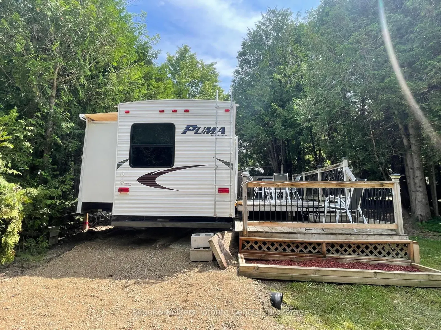 A white Puma RV parked on gravel next to a wooden deck with outdoor furniture, surrounded by green trees.