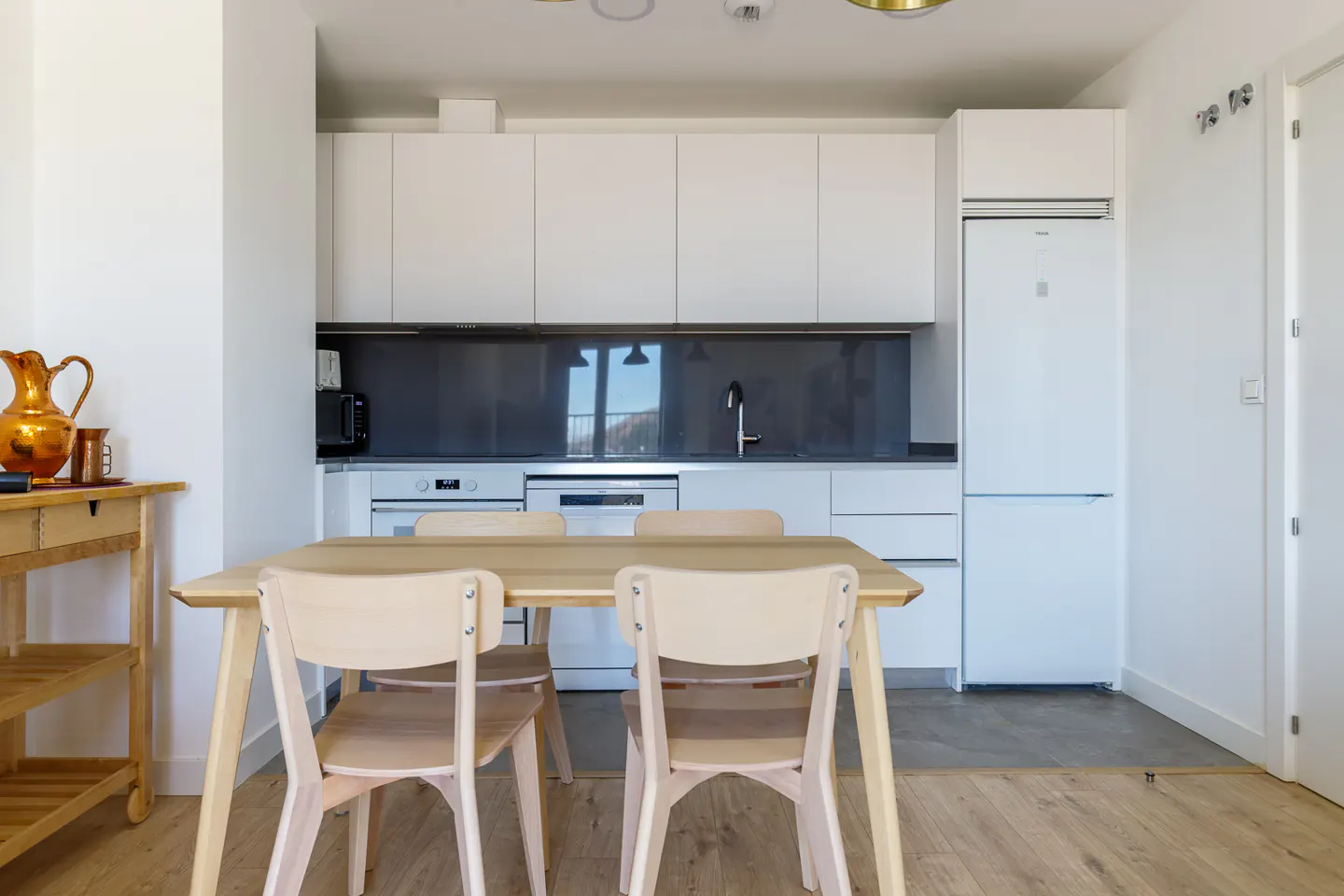 A bright kitchen with white cabinets, a gray backsplash, and a light wood table with four chairs. A copper pitcher sits on a wooden cart.