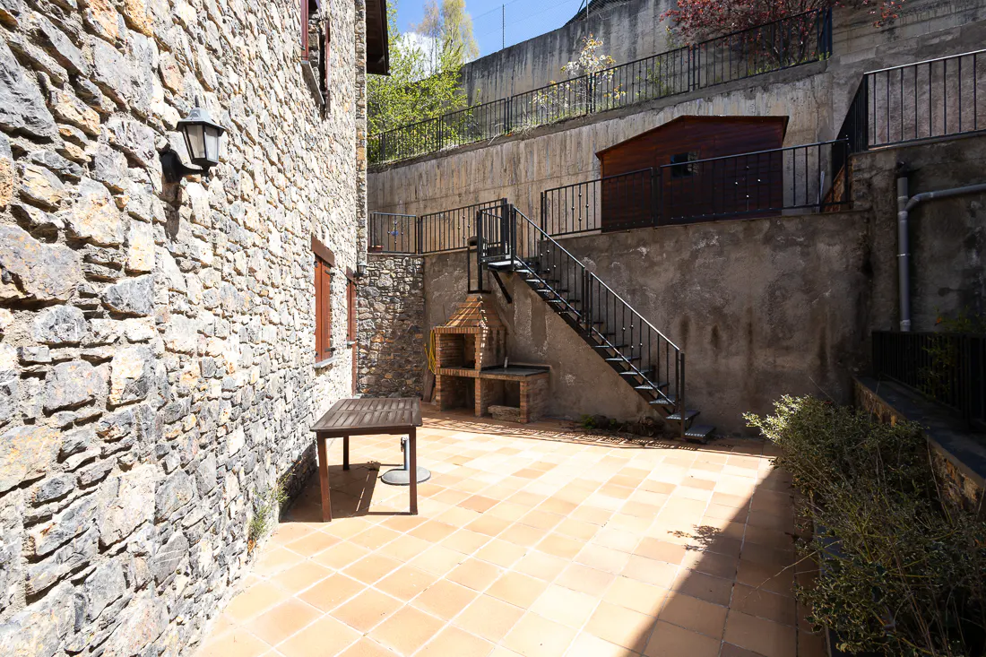 Outdoor patio with stone wall, terracotta tiles, and a dark wood table. A brick grill and metal stairs lead to a shed on a higher level.