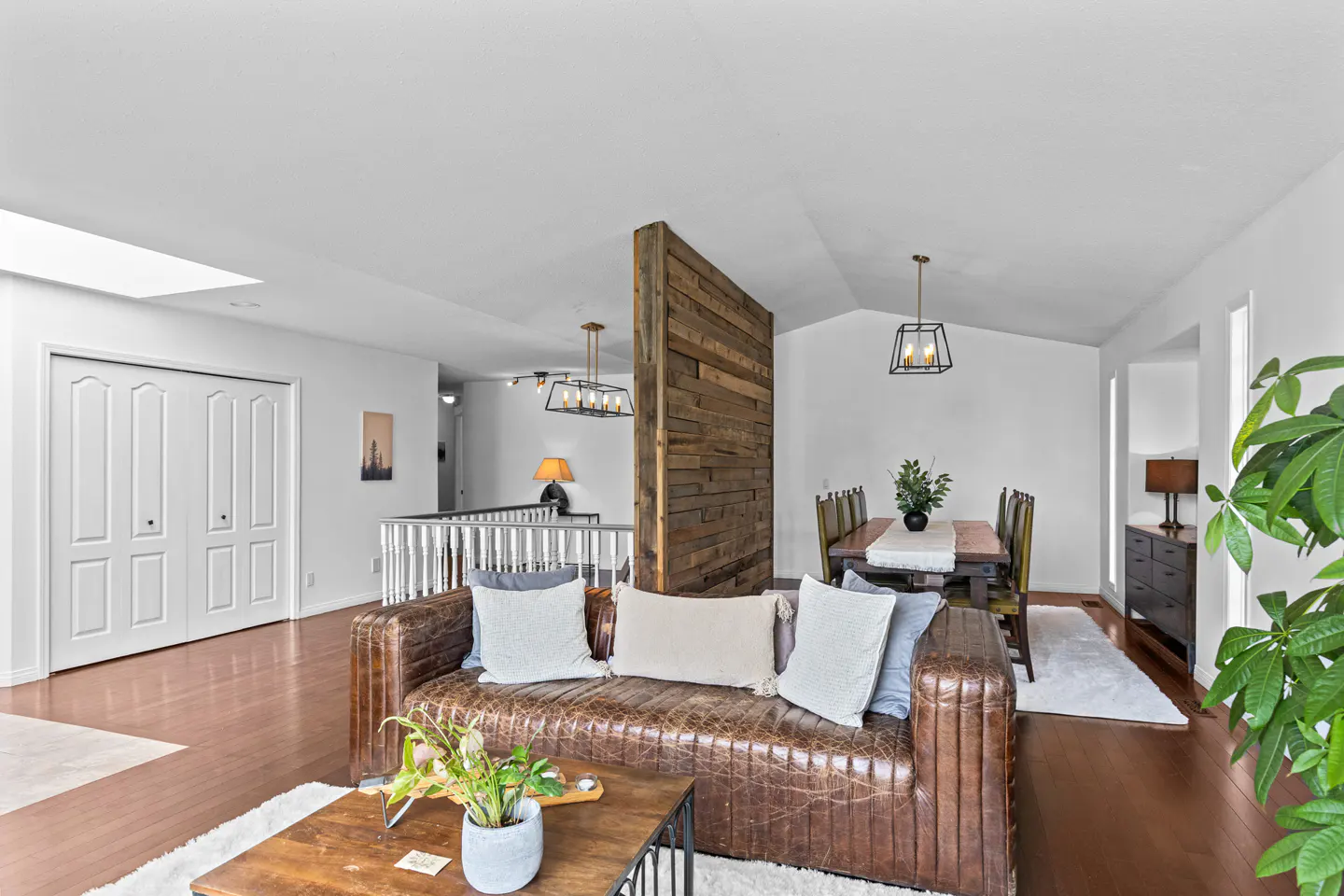 Modern living room with a brown leather sofa, wood accent wall, dining area, and hardwood floors.