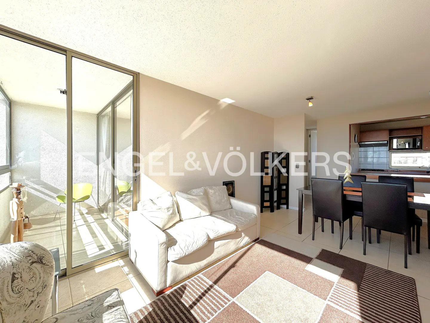 Bright apartment interior with a white sofa, dining table, and balcony with green chairs. A brown and beige rug adds warmth.