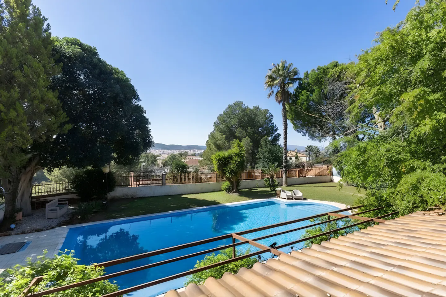 A backyard with a blue pool, green grass, trees, and a view of a city in the distance.