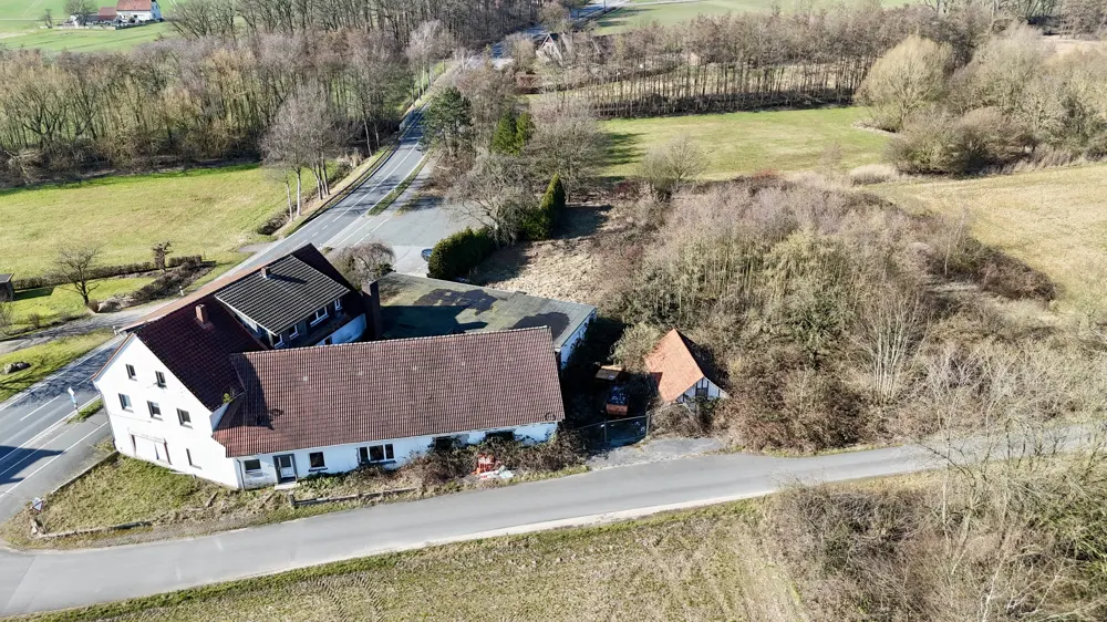 Aerial view of a white farmhouse with a red tile roof, next to a road and surrounded by fields and trees.