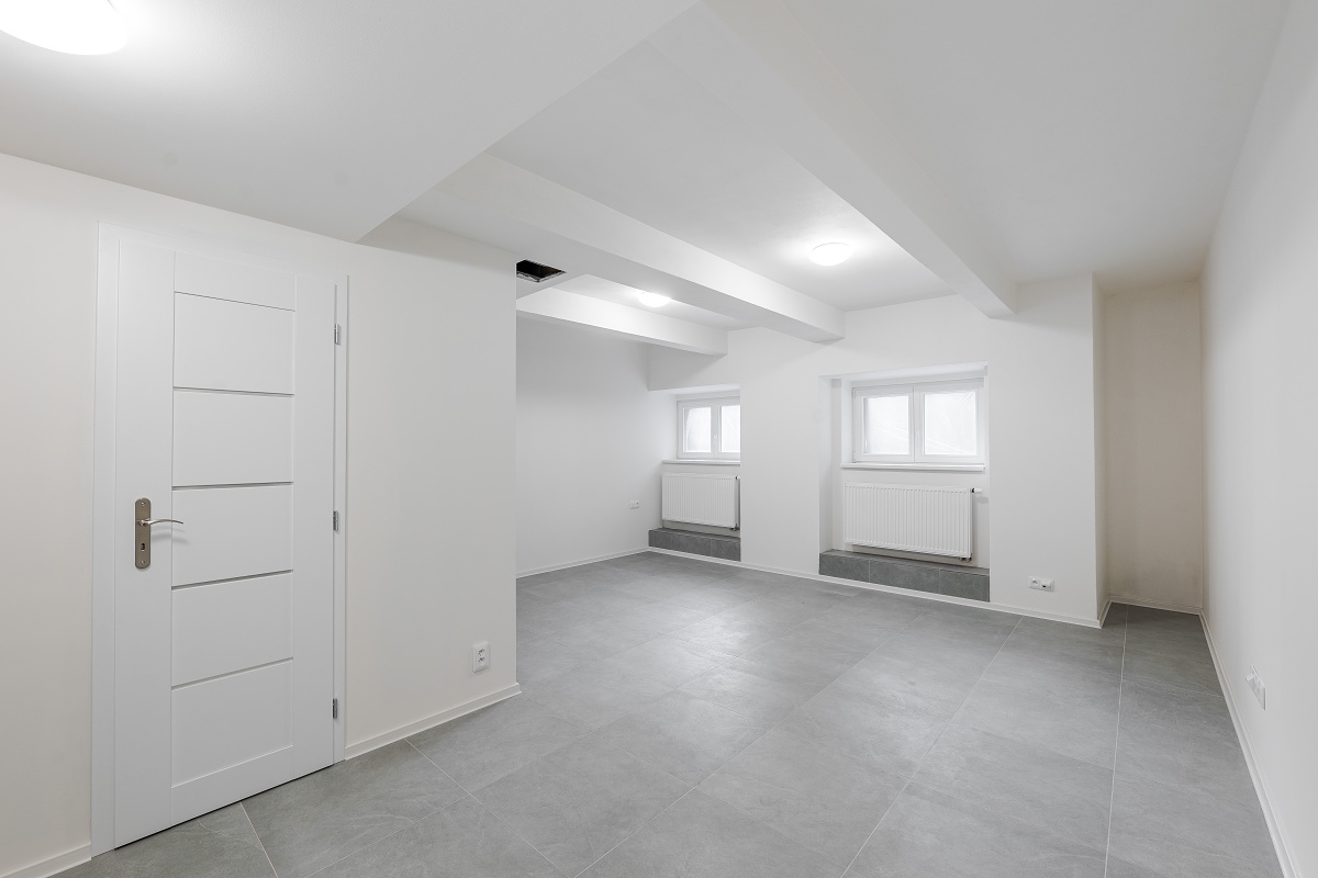 Empty room with white walls, gray tiled floor, a white door, and two windows with radiators under a beamed ceiling.