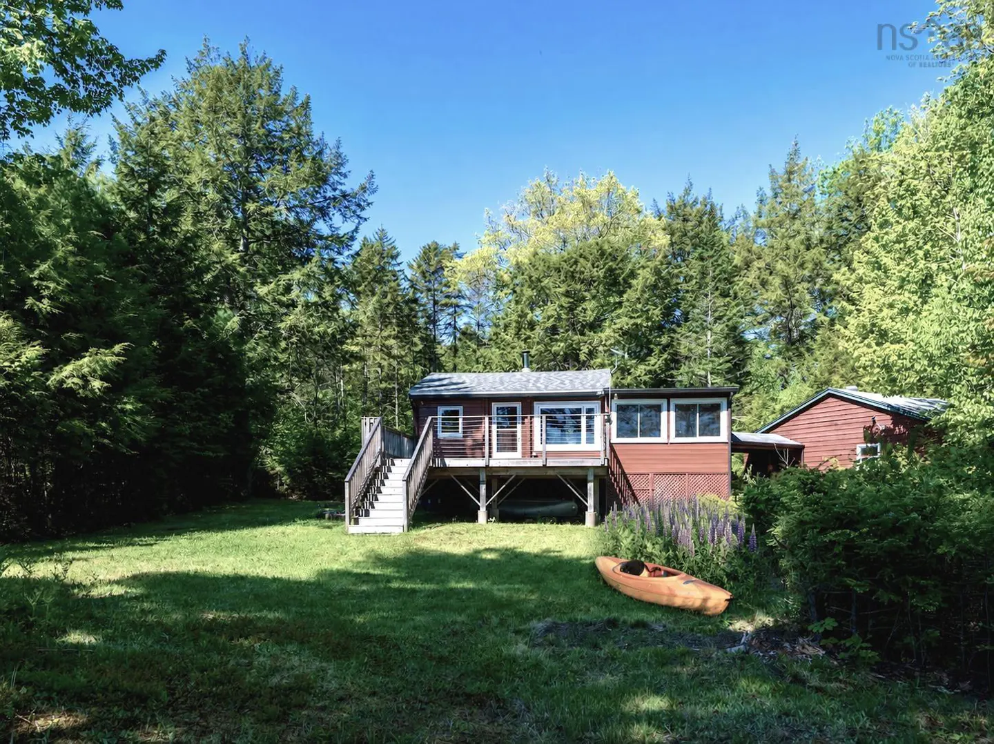 A brown cottage with a raised deck and stairs sits in a lush green yard surrounded by dense forest, with a kayak nearby.