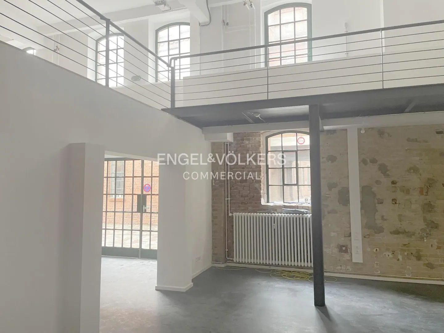 Bright, empty loft with a gray floor, white walls, and large windows. A black metal mezzanine overlooks the space. "Engel & Völkers Commercial" is overlaid.