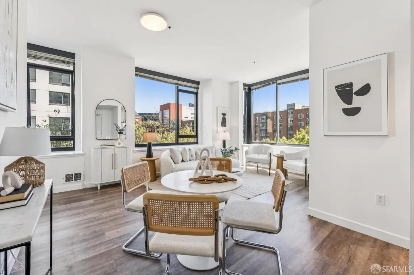 Bright, modern living room with large windows, white walls, and wood floors. A round table with four chairs sits in the foreground. A white sofa and chairs are in the background.