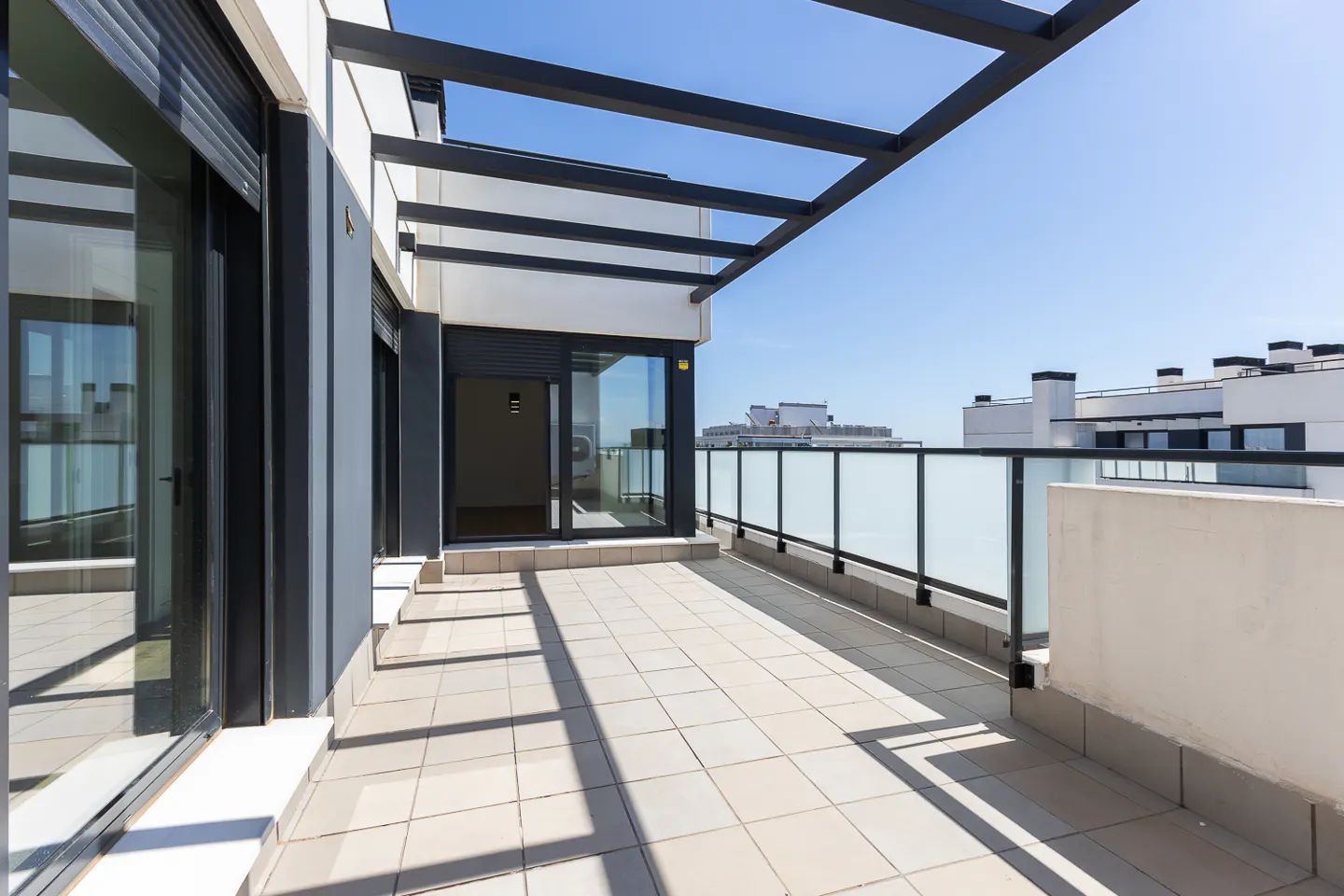 A modern balcony with gray tile flooring, glass railings, and a black metal pergola against a blue sky.
