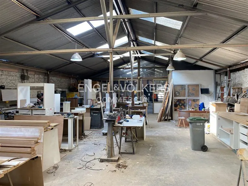 Interior of a large, cluttered workshop with tools, wood, and cabinets under a high, skylit metal roof.