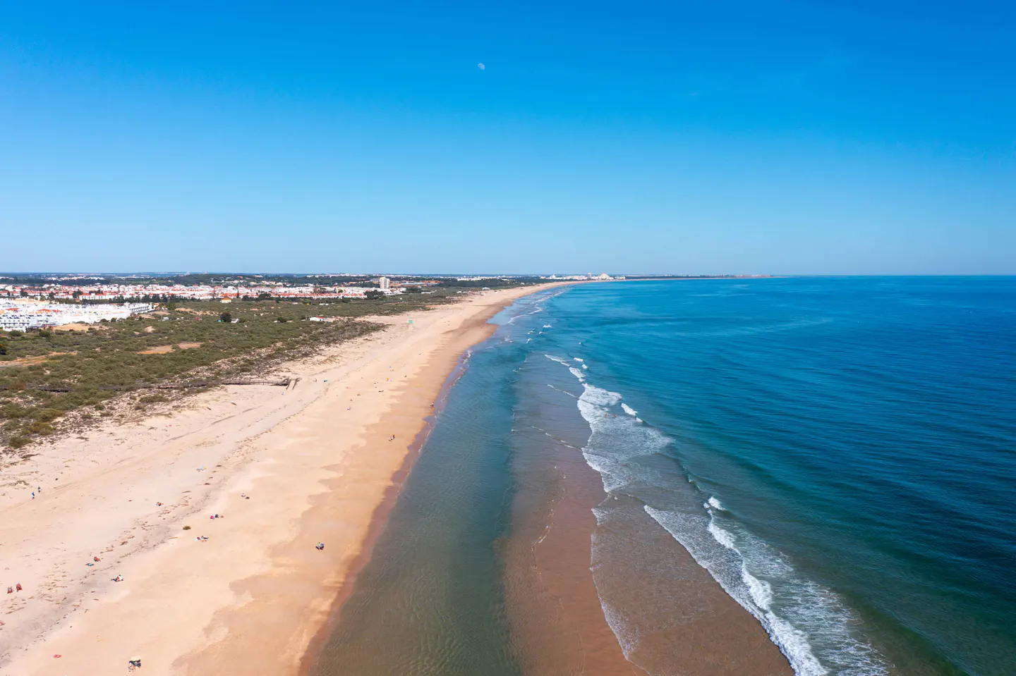 Aerial view of a long, sandy beach meeting the blue ocean under a clear sky. Buildings are visible in the distance.