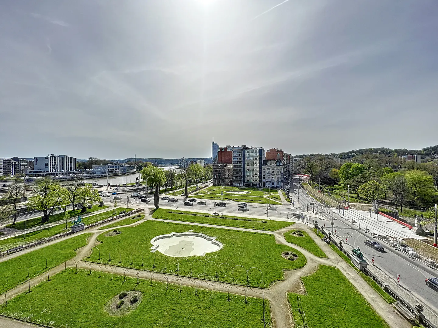 Aerial view of a green park with a dry fountain, buildings, and a river in the background under a cloudy sky.