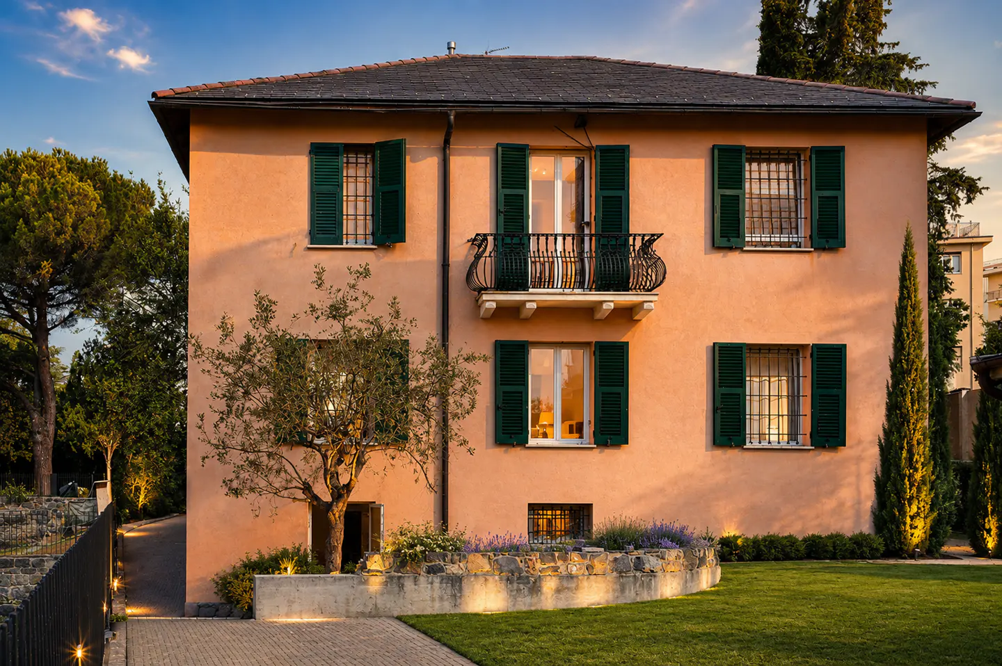 Two-story peach house with green shutters, a black roof, and a small balcony. A tree and green lawn are in front.