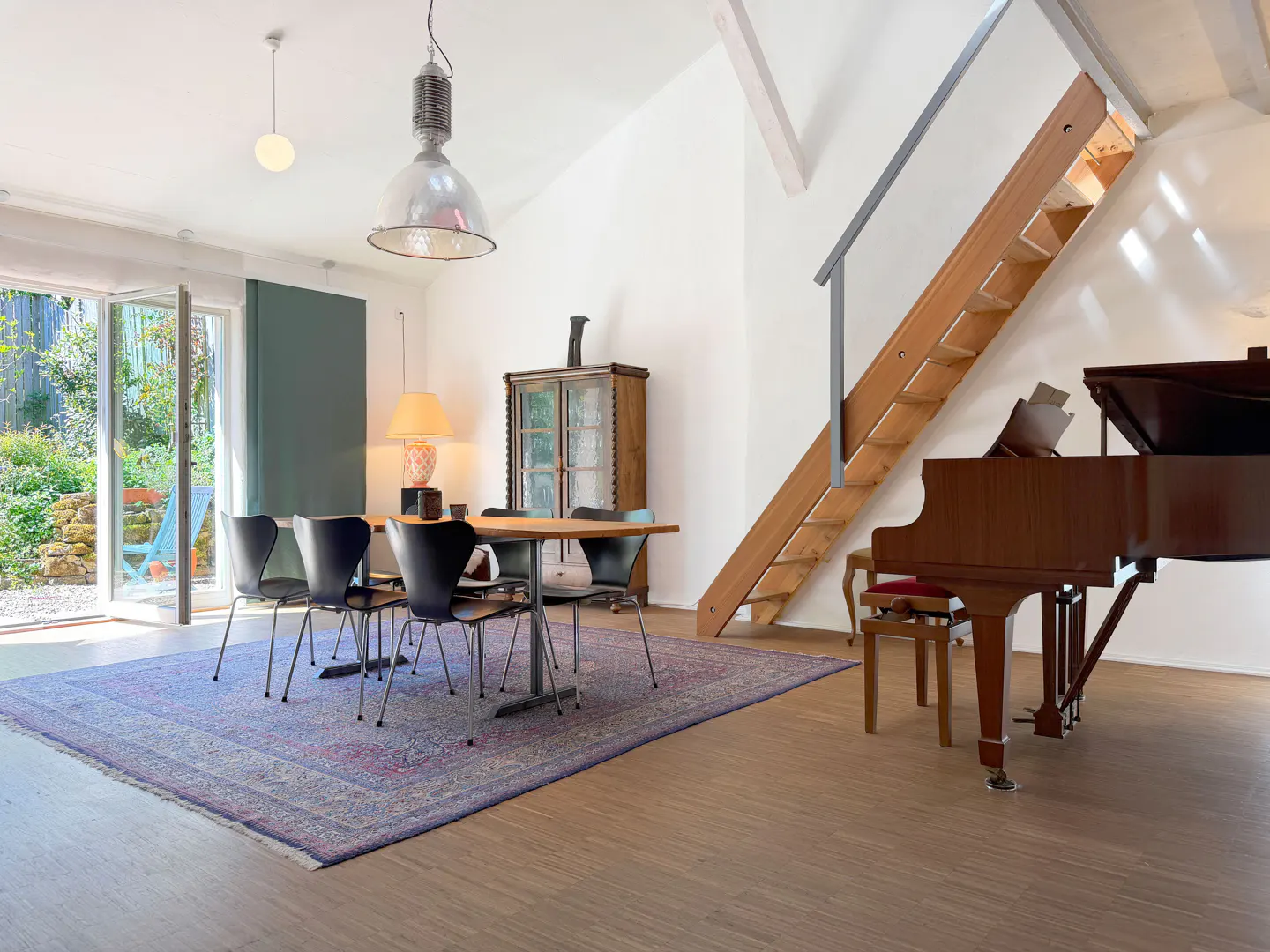 Bright, airy dining room with a wooden table, black chairs, a blue rug, and a grand piano near a wooden staircase.