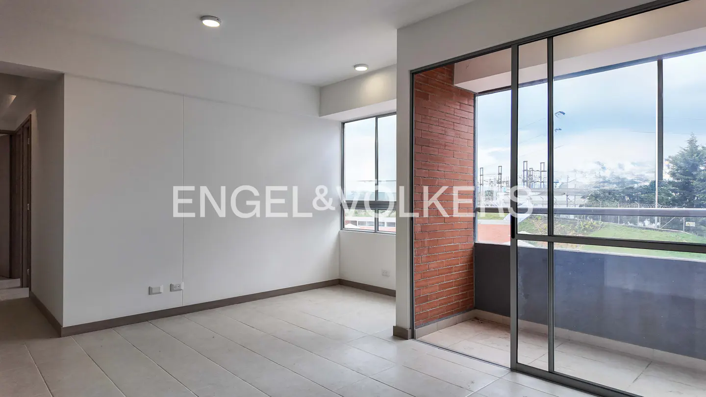 A bright, empty room with white walls, light tile floors, and a brick accent wall leading to a balcony with a view.
