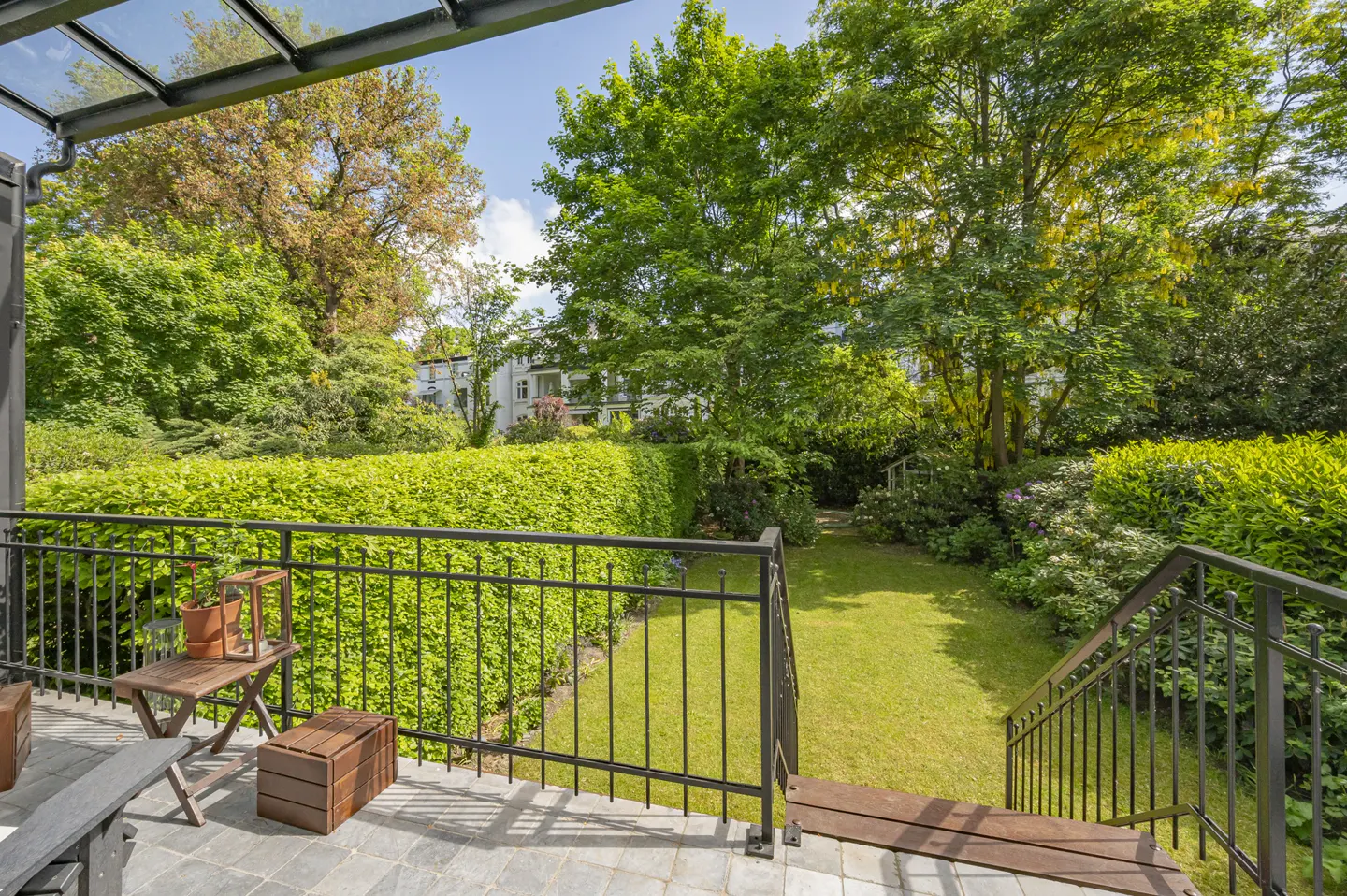 Patio with black metal railing overlooks a green lawn and lush trees. A small table and bench sit on the gray stone patio.