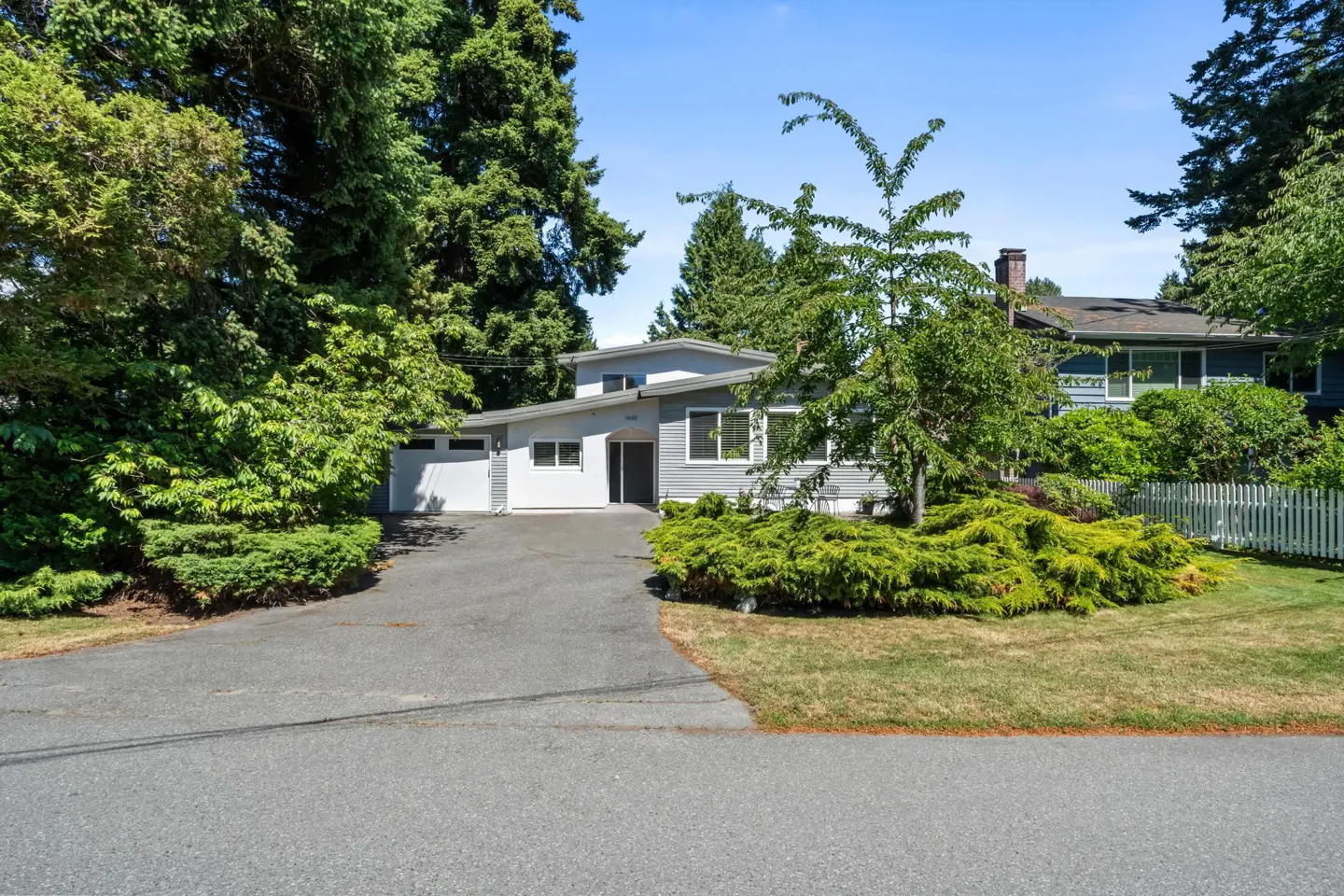 Single-story grey house with a white garage, paved driveway, and lush green landscaping under a clear blue sky.