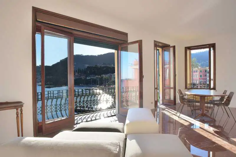 Bright living room with marble floors, a white sofa, and a table with chairs. Open doors lead to a balcony with a sea view.