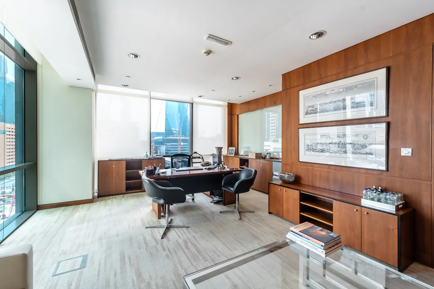 A modern office with a large desk, black chairs, wood paneling, and a city view from the window.