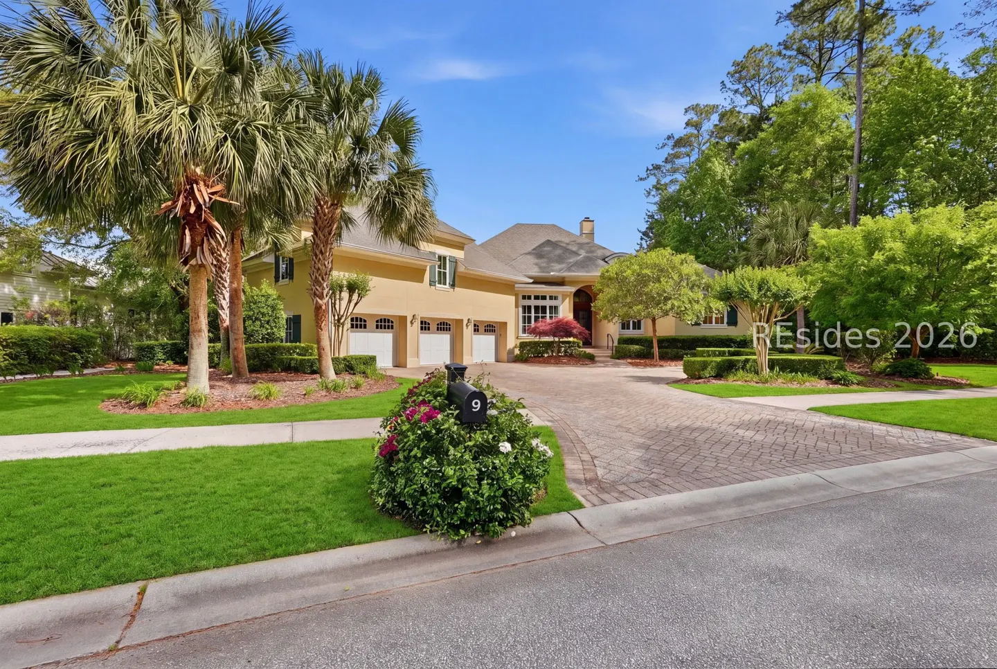 A tan two-story house with a three-car garage, palm trees, and a brick driveway on a sunny day.