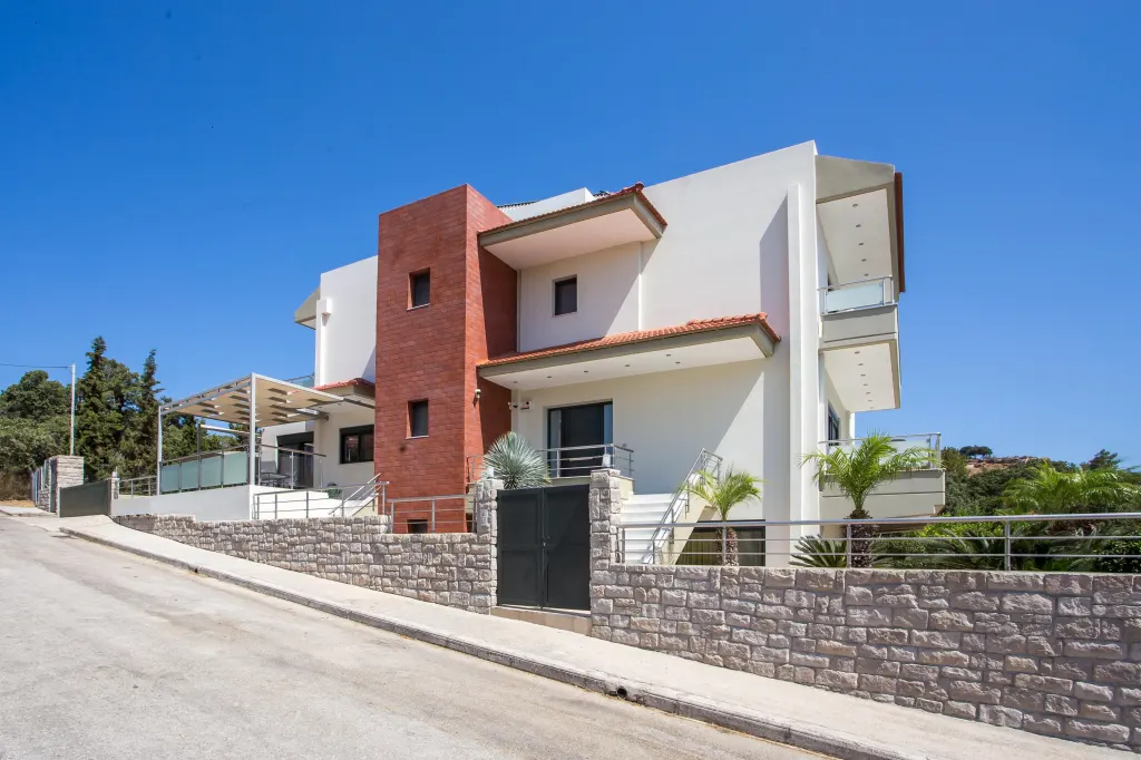 Modern white house with red brick accent, balconies, and a stone wall under a clear blue sky.