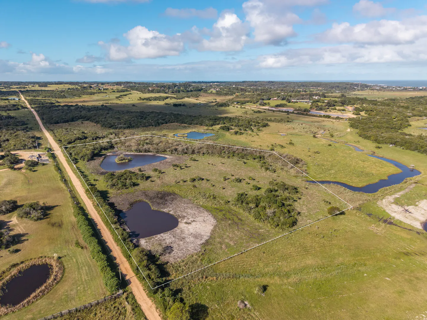 Aerial view of a large, green, grassy property with ponds, trees, and a dirt road, outlined in white. Blue sky with clouds.