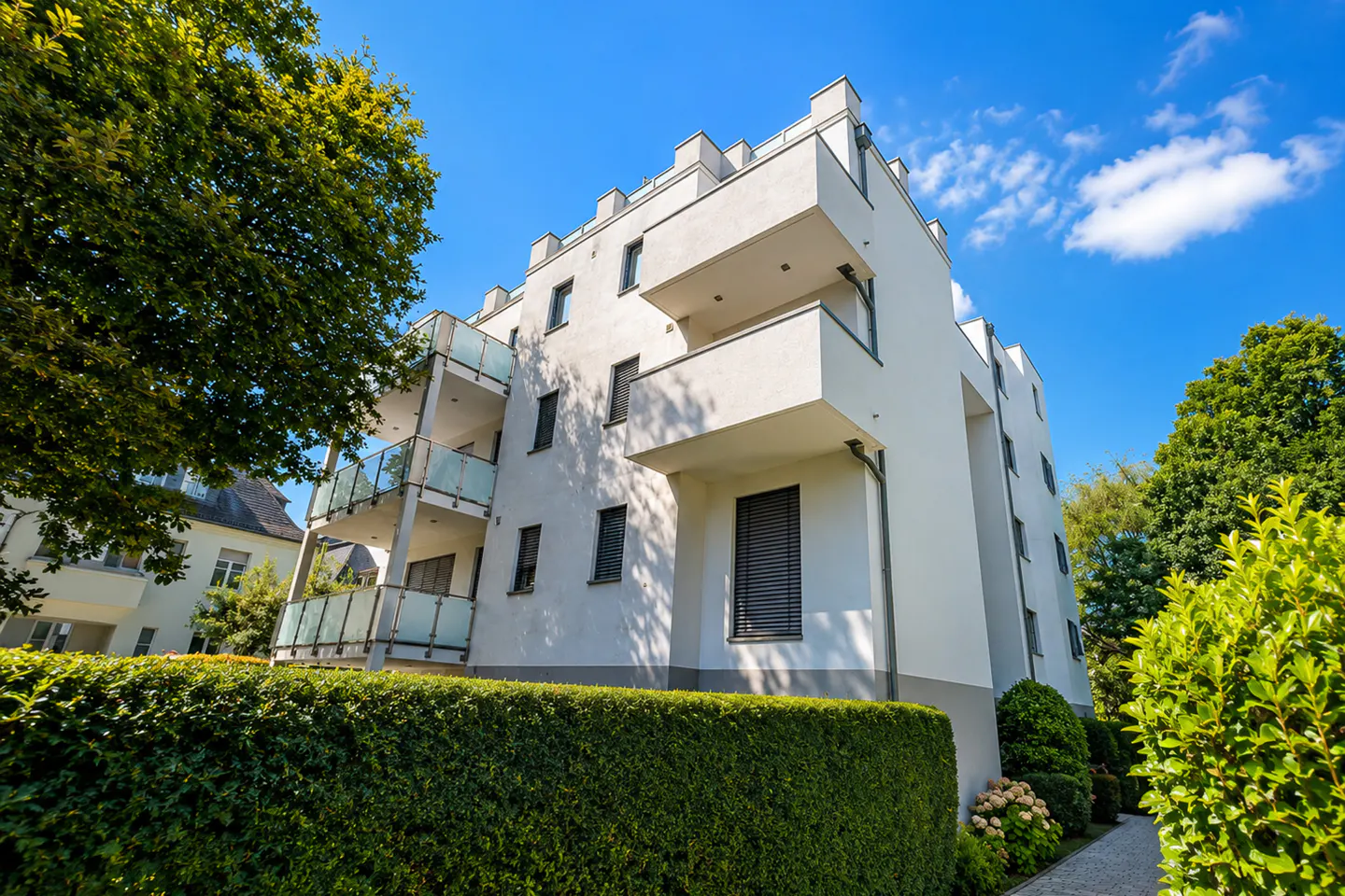 Modern white apartment building with balconies, set against a blue sky and green trees.
