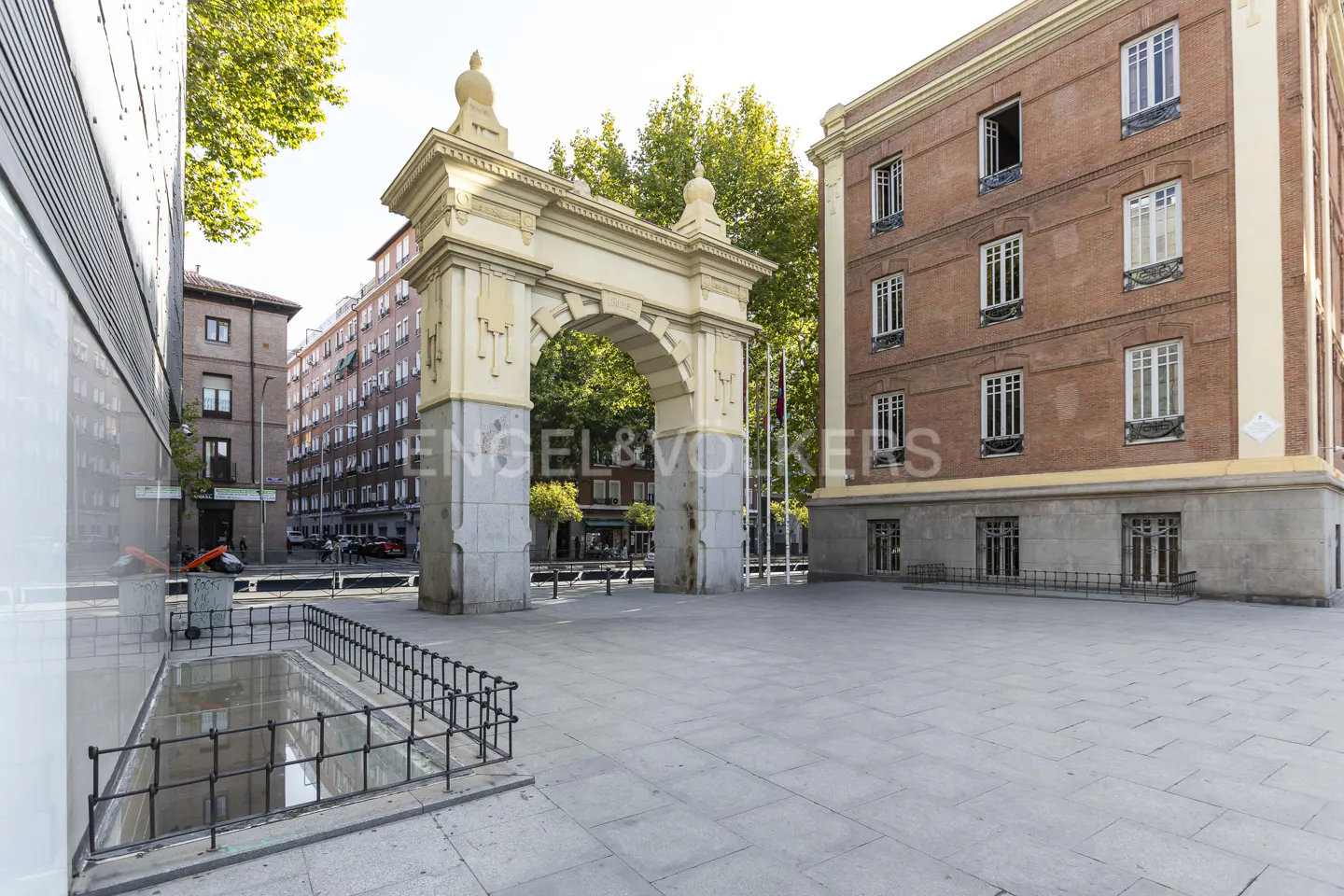 A wide shot of a plaza with a stone archway, brick buildings, and trees.