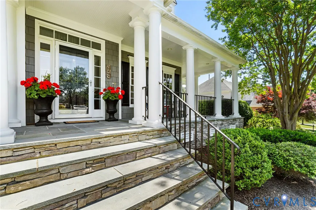 Stone steps lead to a porch with white columns. Red flowers sit in black urns by the front door.