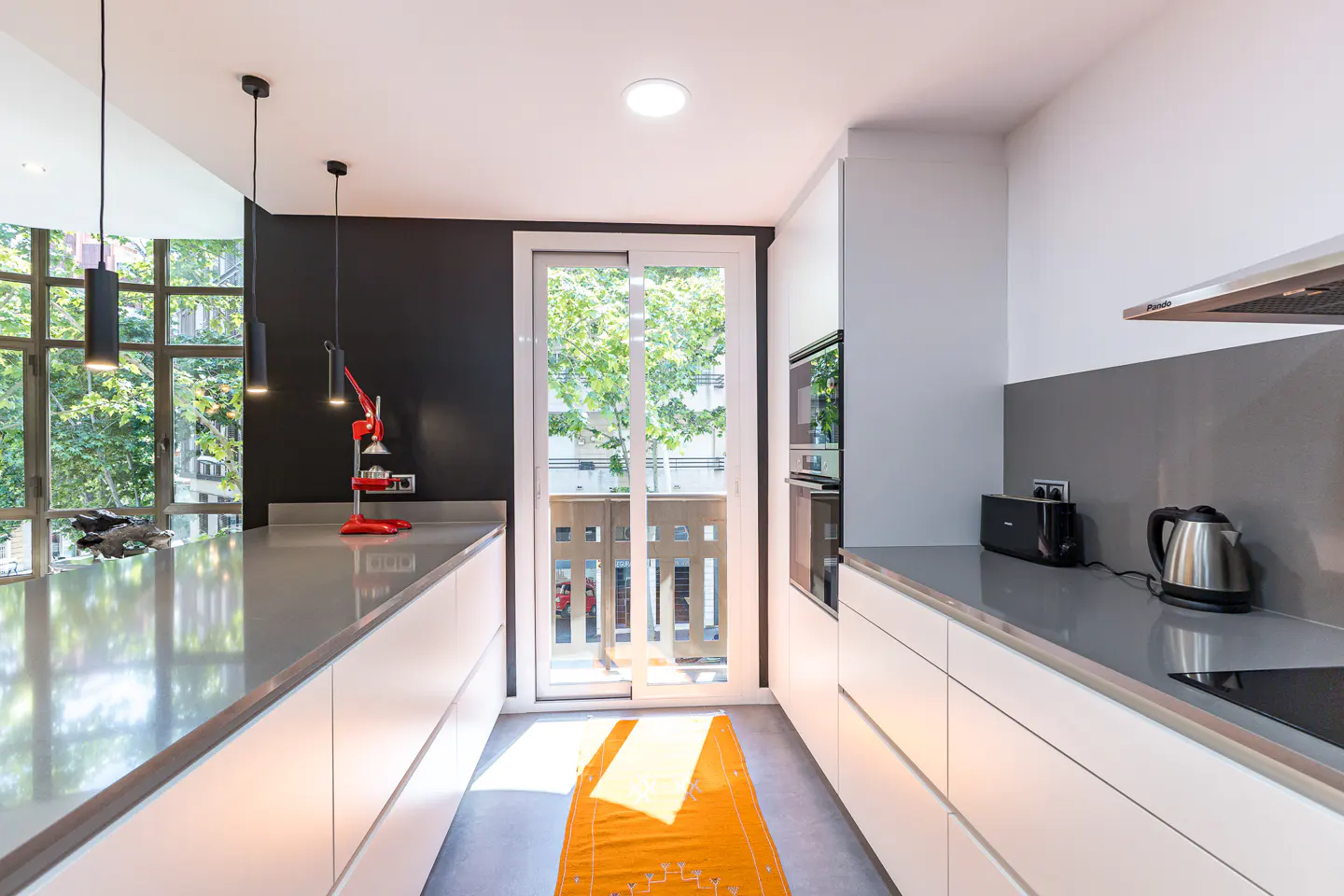 A modern kitchen with white cabinets, gray countertops, and black accent wall. A red juicer sits on the counter near a window and sliding glass door.