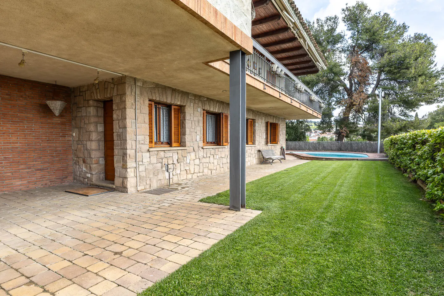 Exterior view of a stone house with a green lawn, pool, and trees in the background. The house has wooden shutters and a balcony.