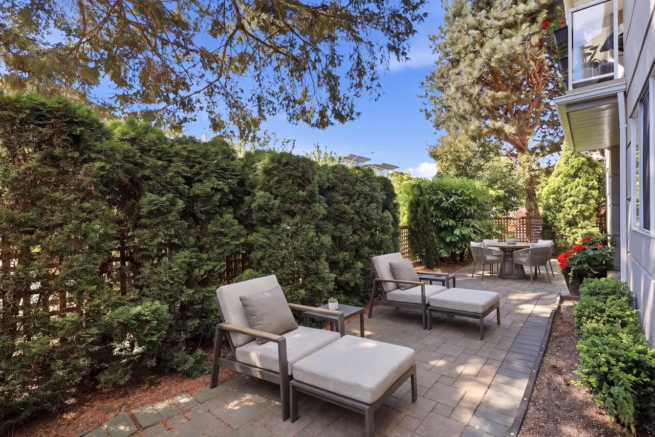 A backyard patio with two lounge chairs, a small table, and a dining set, surrounded by green bushes and trees.