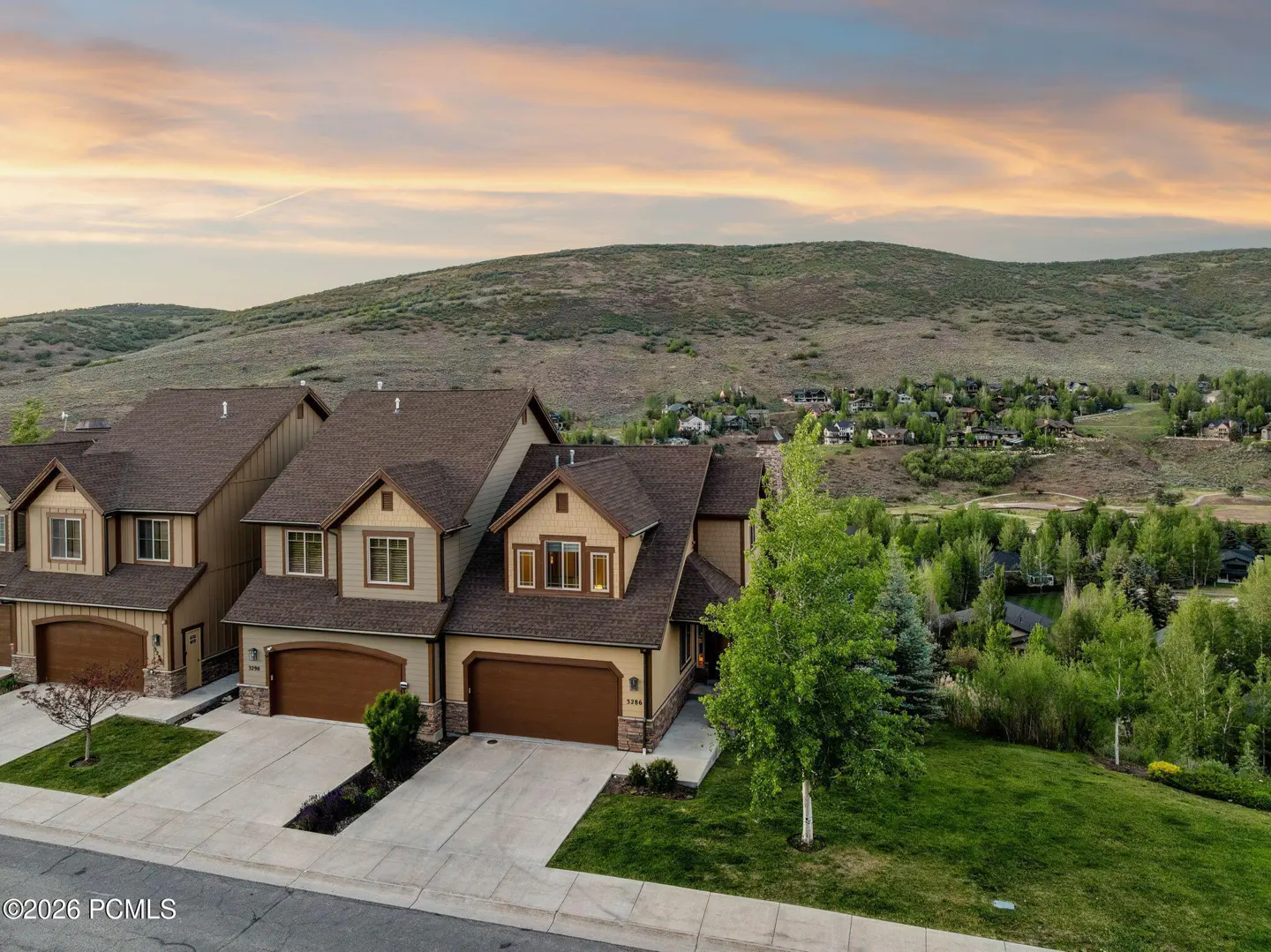 Two-story tan townhomes with brown roofs and garages, set against a scenic mountain landscape at sunset.