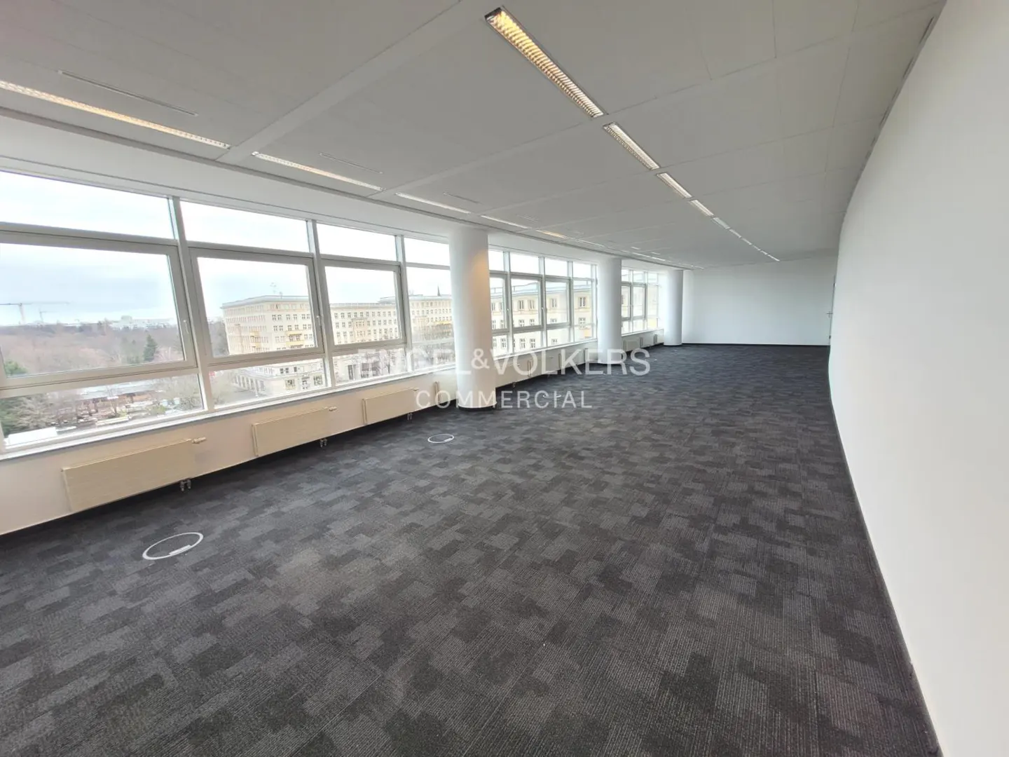 Bright, empty office space with gray carpet, white walls, and large windows overlooking a city.