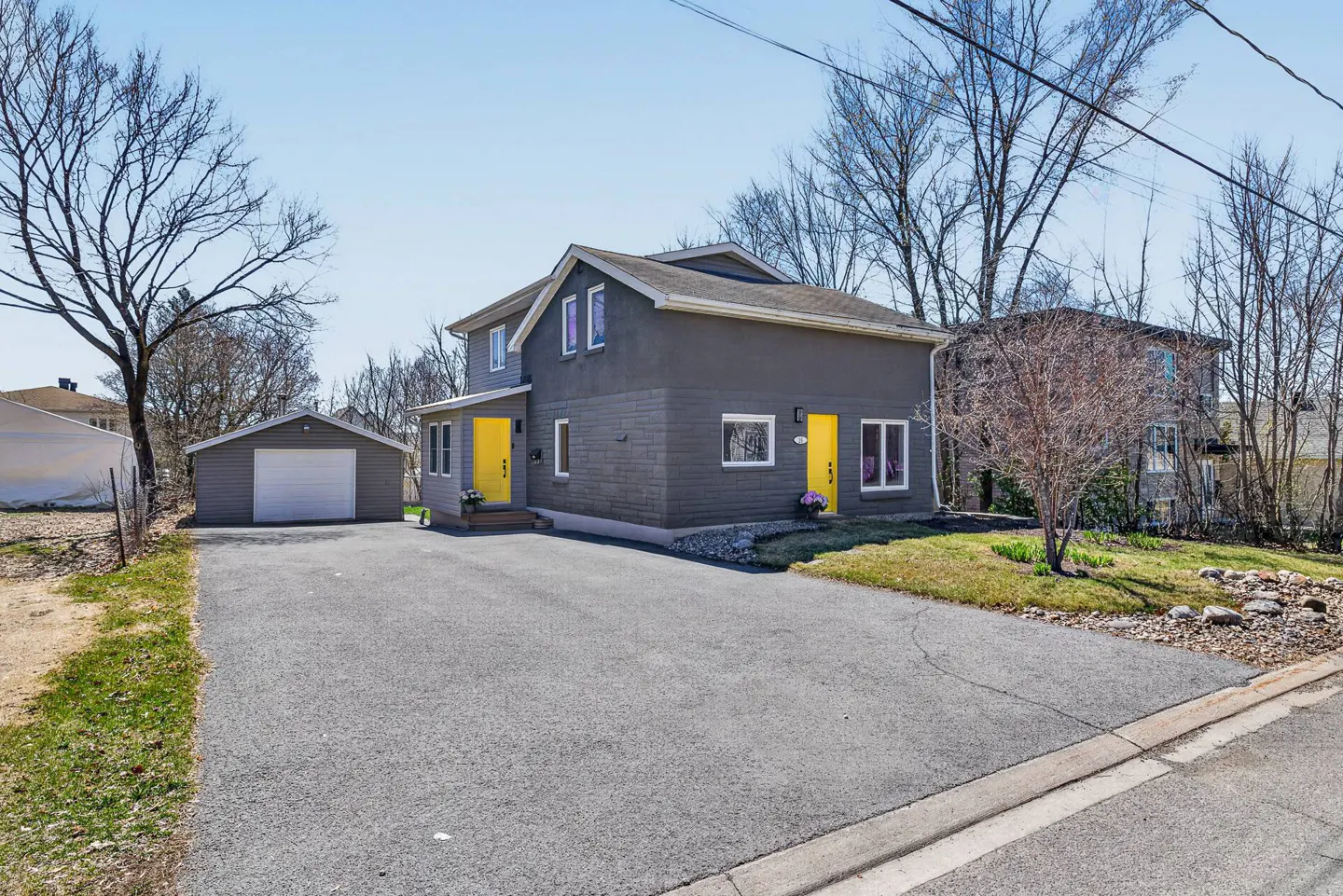 Two-story grey house with two bright yellow doors, a detached garage, and a paved driveway under a clear blue sky.
