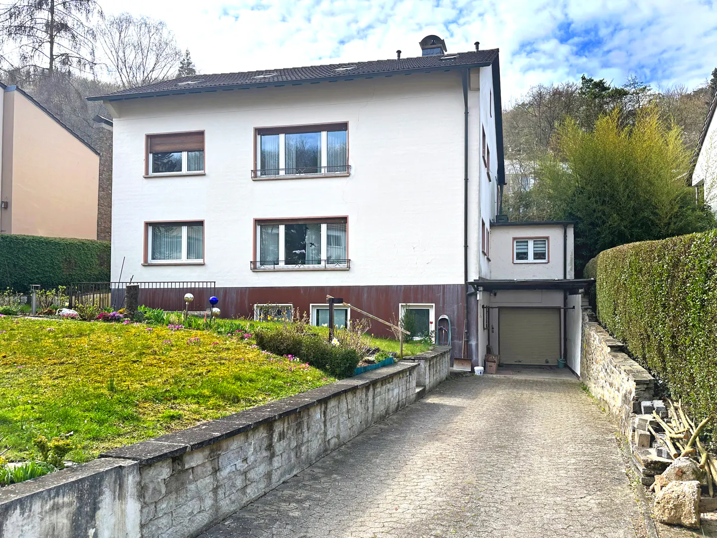 Two-story white house with a brown base, a garage, and a stone driveway leading up to it. Green lawn and trees surround the house.
