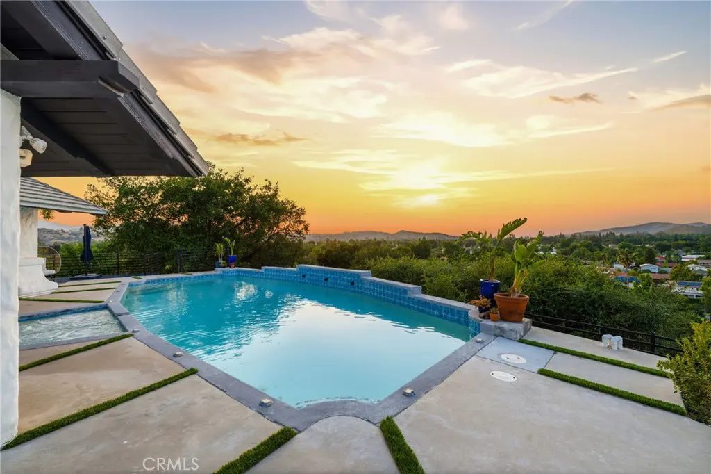Outdoor pool with blue tiles and concrete deck overlooking a sunset view of trees and distant hills.