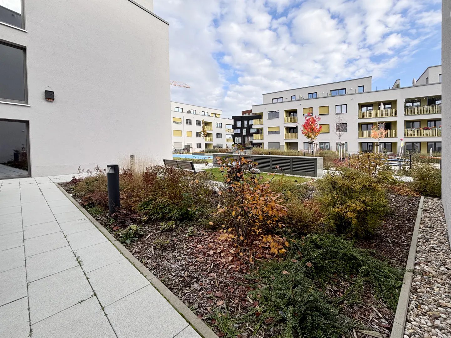 Modern apartment complex courtyard with paved walkways, landscaped garden beds, and autumn foliage under a blue sky.