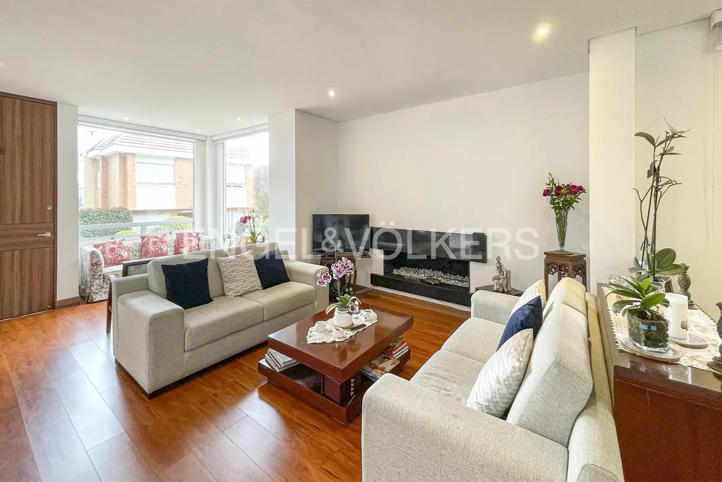 Living room with hardwood floors, two beige sofas, a dark wood coffee table, and a fireplace with a TV above it.