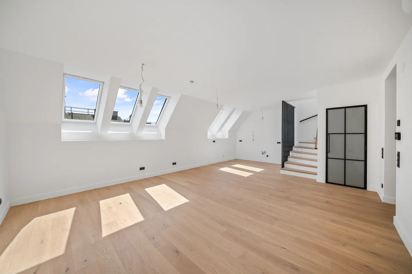 Bright, empty attic room with white walls, light wood floors, and skylight windows. Stairs and a black-framed glass door are visible.