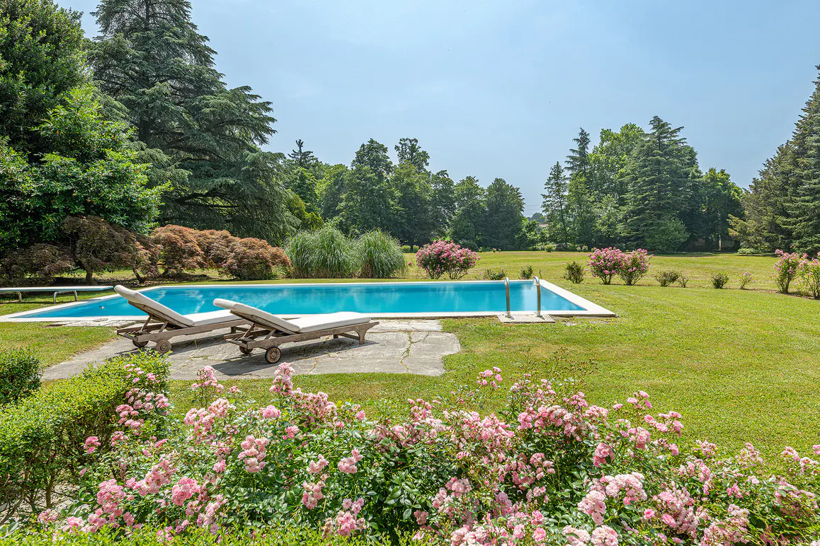 A rectangular pool with blue water is surrounded by green grass, trees, and pink roses. Two lounge chairs sit on the pool deck.