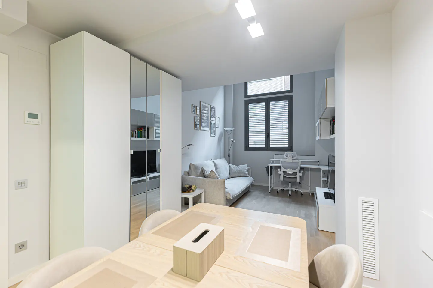 A bright, modern studio apartment with a dining table in the foreground and a living area with a sofa and desk in the background.