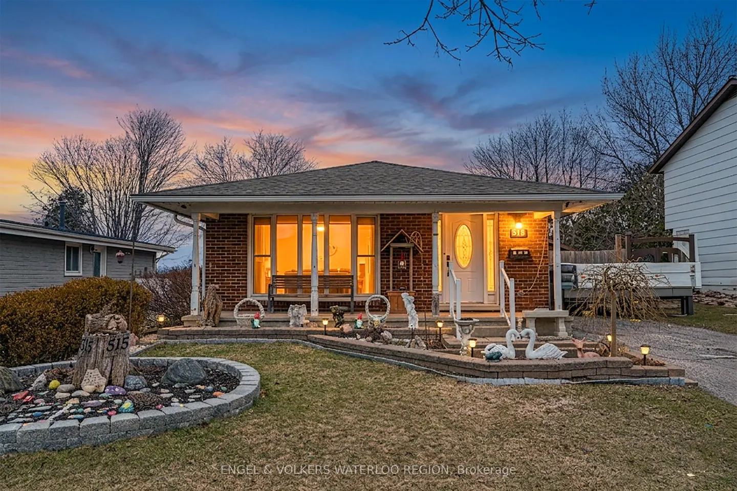 A single-story brick house with a white door and porch at dusk. The lawn has decorative elements.